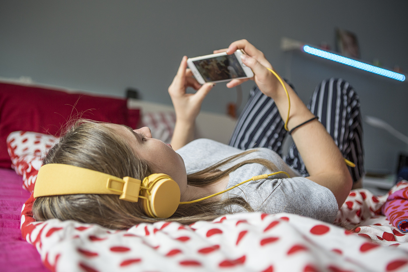 Un'adolescente sdraiata su un letto che guarda qualcosa con le cuffie del suo smartphone.