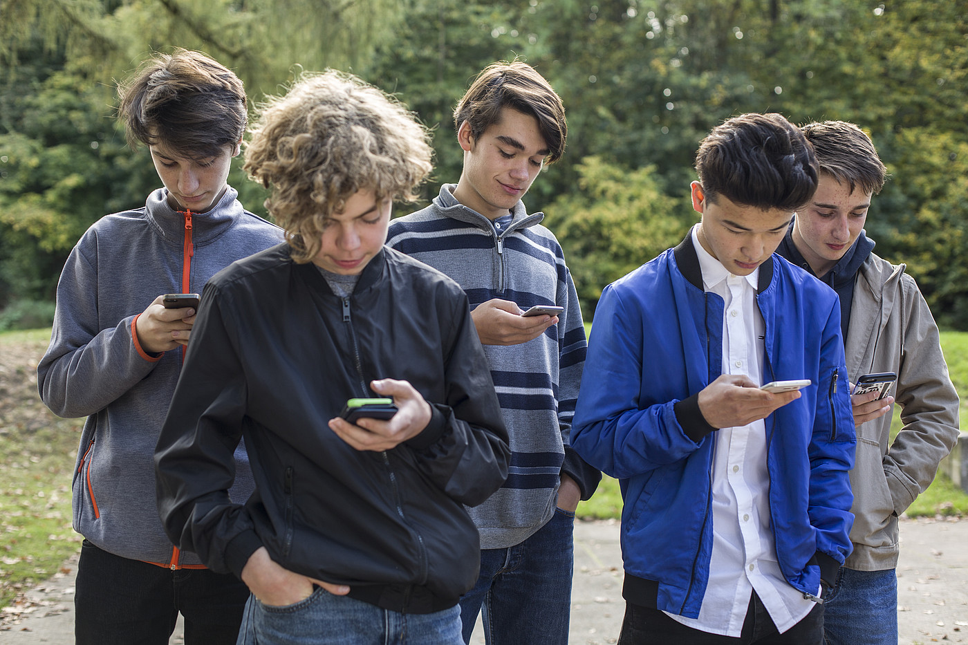Cinque adolescenti in piedi fuori e tutti a guardare i loro smartphone.