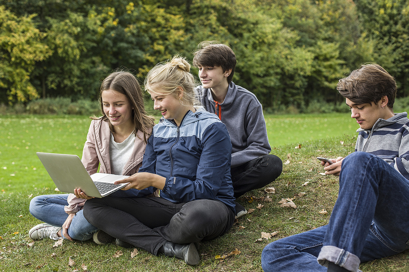 Quattro adolescenti seduti su un prato. Tre di loro guardano insieme un computer portatile, una persona uno smartphone.