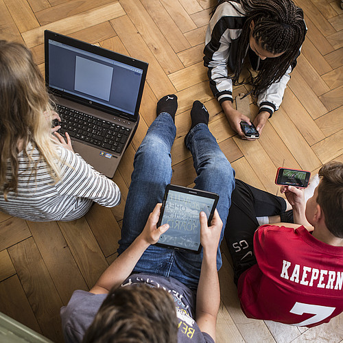 4 adolescenti seduti o sdraiati sul pavimento, tutti con un computer portatile, un tablet o uno smartphone.