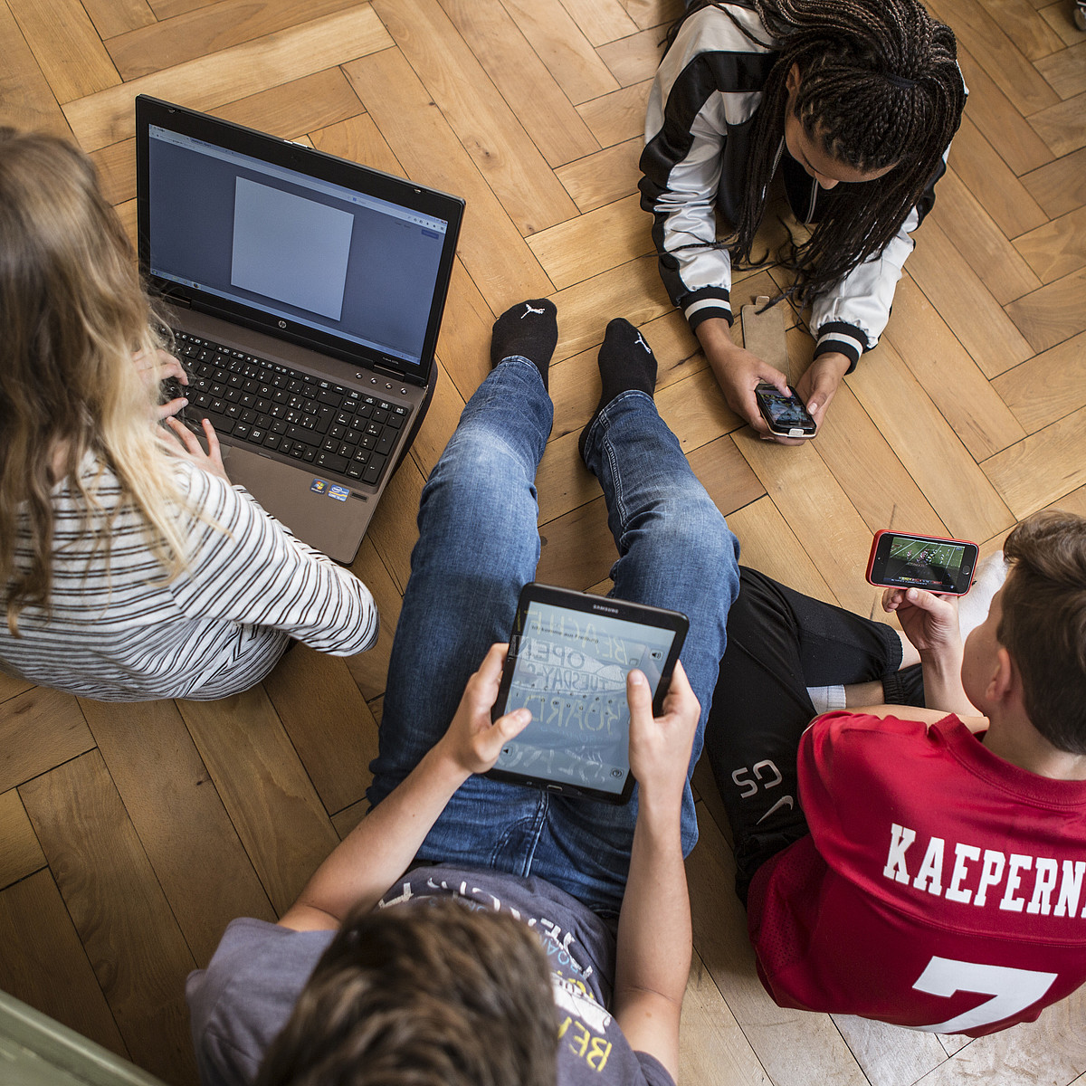 4 adolescenti seduti o sdraiati sul pavimento, tutti con un computer portatile, un tablet o uno smartphone.