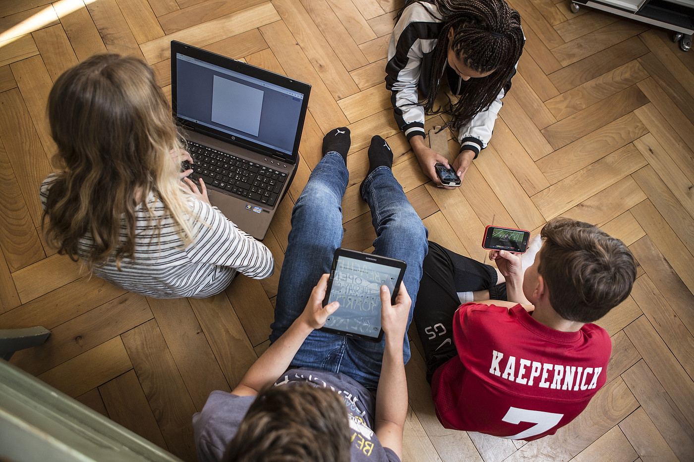 4 adolescenti seduti o sdraiati sul pavimento, tutti con un computer portatile, un tablet o uno smartphone.
