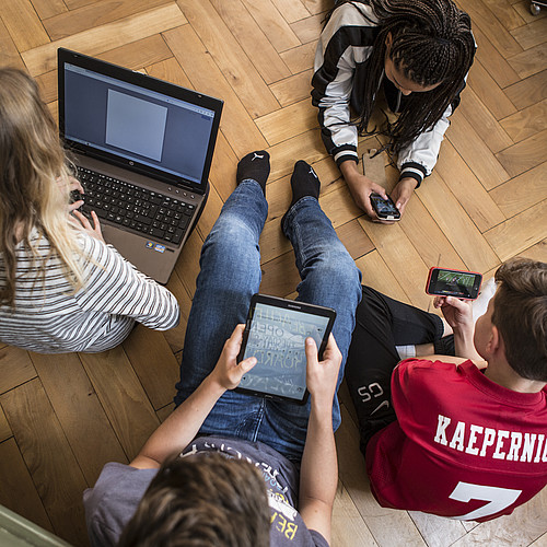 4 adolescenti seduti o sdraiati sul pavimento, tutti con un computer portatile, un tablet o uno smartphone.