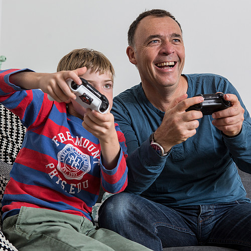 Un padre seduto sul divano con suo figlio che gioca ai videogiochi. 