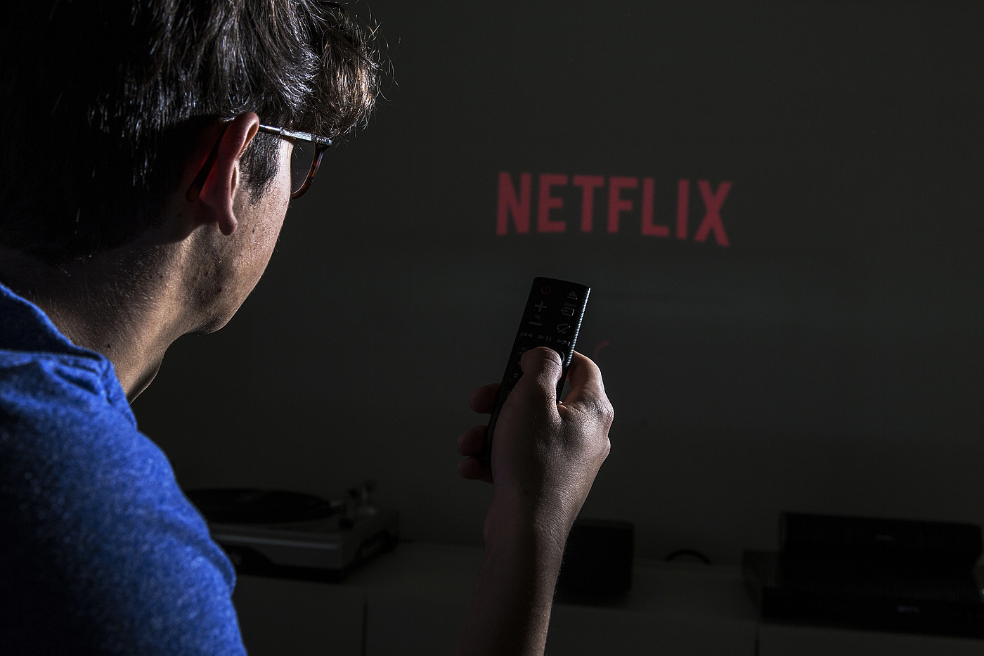 Una persona con un telecomando in mano. Sullo sfondo si può vedere il logo di Netflix.