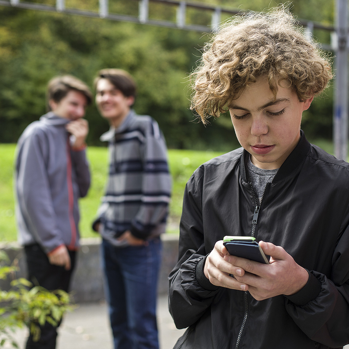 Un giovane è fuori con il suo cellulare. Sullo sfondo ci sono altri due giovani che ridono di lui.