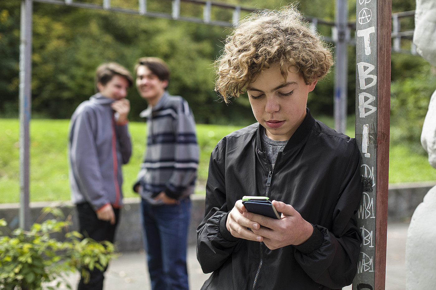 Un giovane è fuori con il suo cellulare. Sullo sfondo ci sono altri due giovani che ridono di lui.