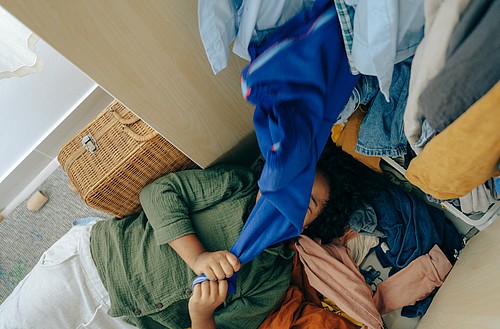 [Translate to Italienisch:] Frau liegt vor einem Schrank unter Kleidern begraben.
